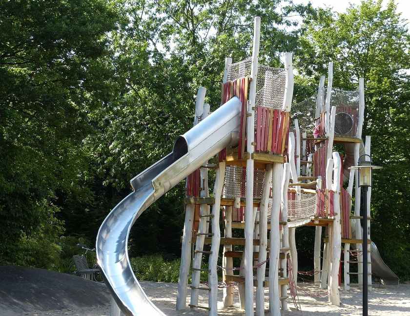 Structure de jeu avec toboggan en métal, poutres en bois, filet, dans un parc arboré. Enfants jouent