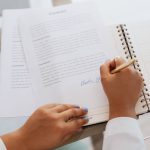 Une main signe un document dans un carnet spiralé, devant un ordinateur portable