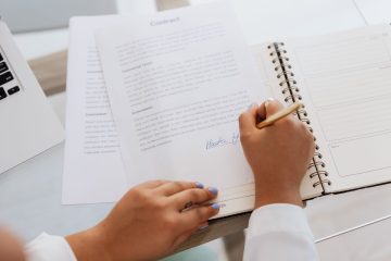 Une main signe un document dans un carnet spiralé, devant un ordinateur portable