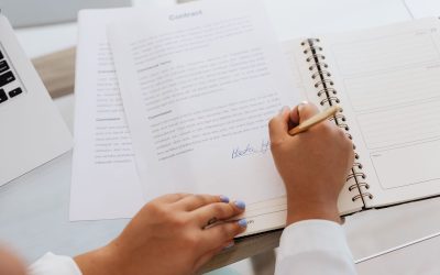 Une main signe un document dans un carnet spiralé, devant un ordinateur portable